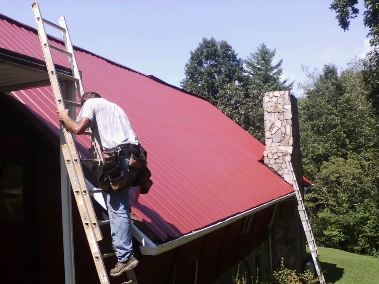 Roofer on a ladder beside a red metal roof and stone chimney — Soffit & Fascia Repair by Roofing Brothers in Lakeview